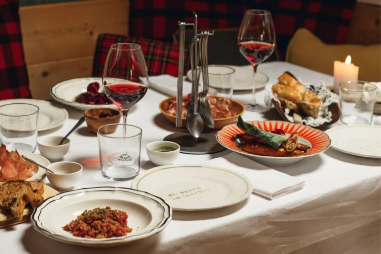 Tavola del Brite de Larieto con piatti decorati, calici, posate al centro e tessili a quadri sullo sfondo